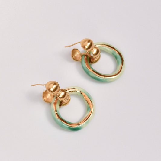 Handmade porcelain hoop earrings featuring a unique circular design with a glossy green and gold enamel finish. These artisanal ceramic earrings are mounted on gold-toned studs with a minimalist yet bold aesthetic. The combination of smooth porcelain and shimmering enamel creates an eye-catching, modern statement piece, perfect for artistic, boho, or contemporary fashion styles. Ideal for everyday wear or special occasions, these earrings reflect a handcrafted, elegant charm and are suitable for gift-giving