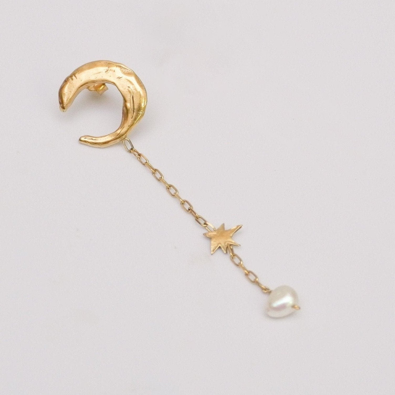 Asymmetric handmade artisanal drop stud earring with a gold-colored organic moon shape, connected to a chain with a star shape, and ending with a small natural pearl.